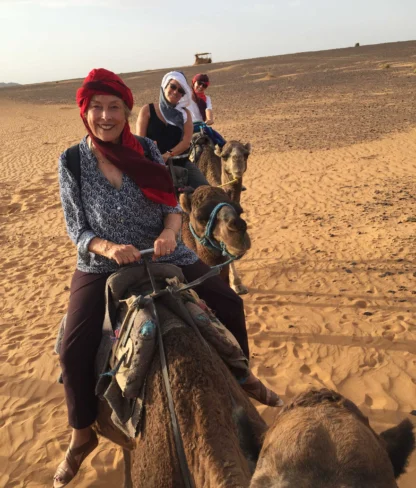 Three people riding camels in a desert landscape, wearing headscarves and casual attire under a clear sky.