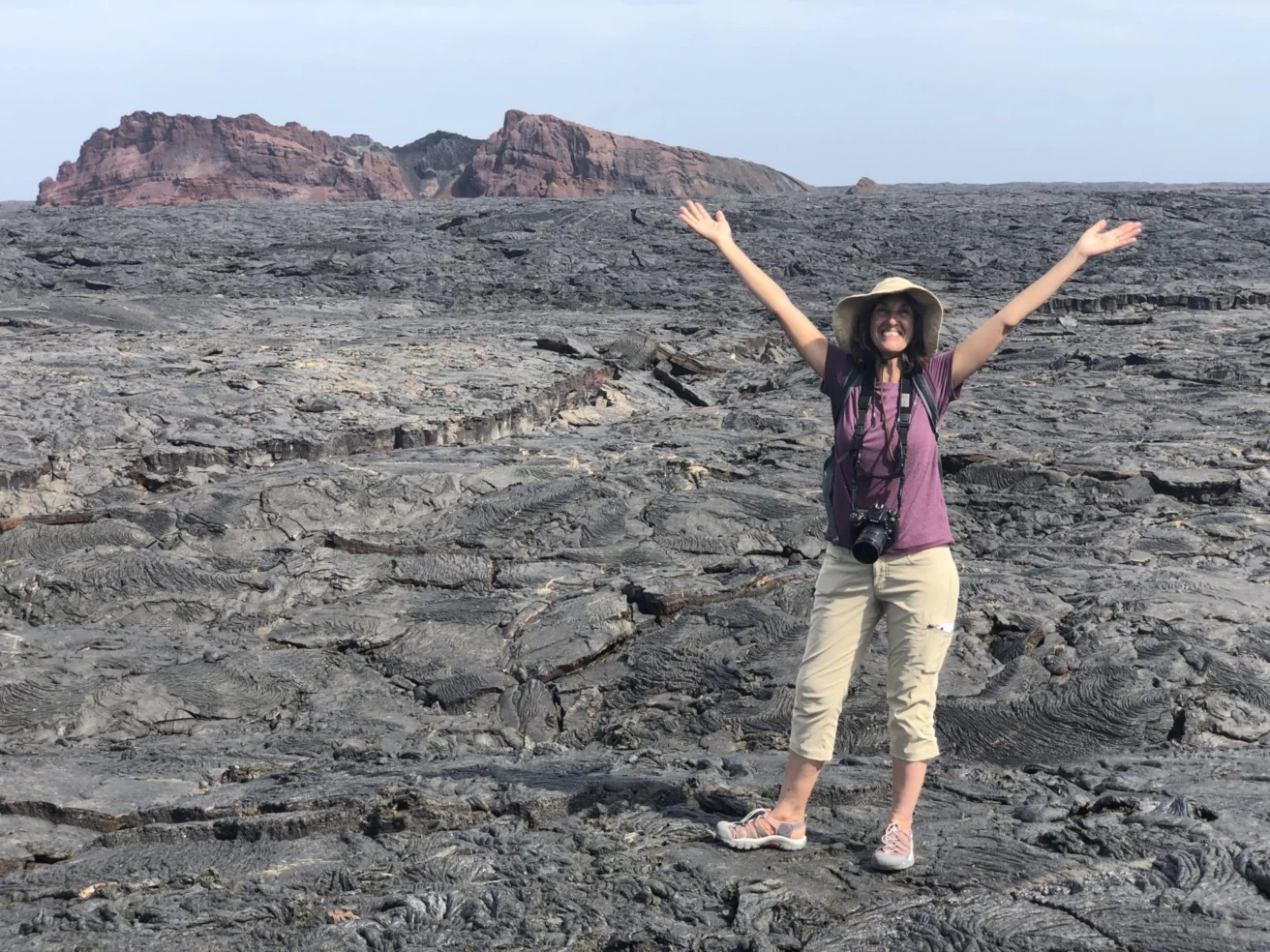 A person stands on a vast lava field with arms raised. A distant rocky formation is visible under a clear sky.