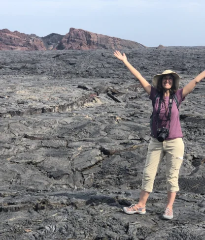 A person stands on a vast lava field with arms raised. A distant rocky formation is visible under a clear sky.