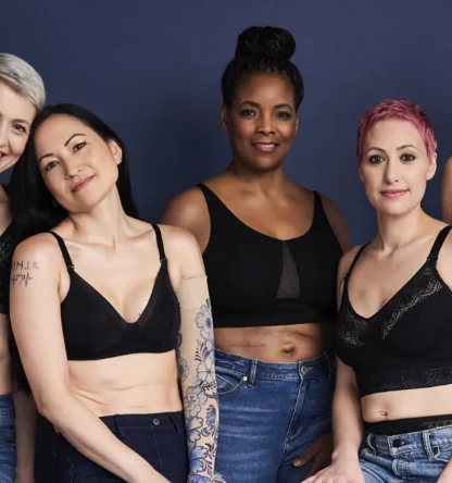 Five women in black bras and jeans stand against a dark blue background, smiling at the camera.