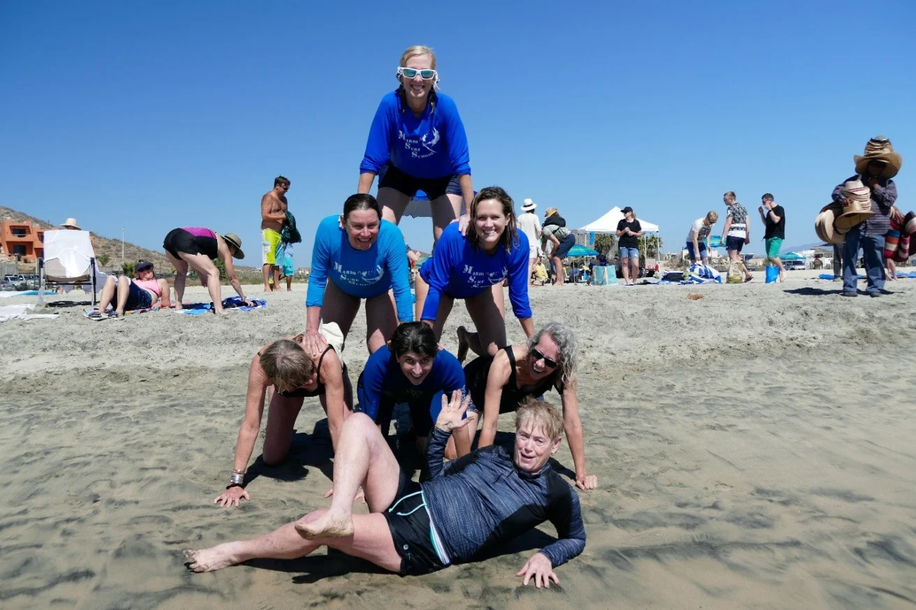 A group of people making a human pyramid on a beach, with a person lying in front of them on the sand, under a clear blue sky.