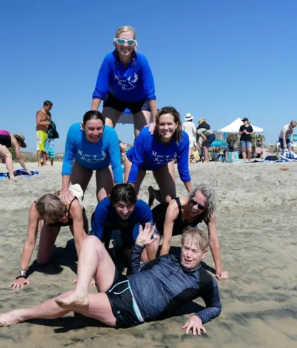 A group of people making a human pyramid on a beach, with a person lying in front of them on the sand, under a clear blue sky.