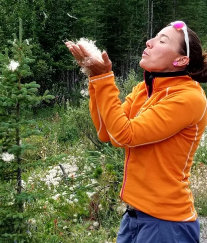 A person in an orange jacket blows white seeds from their hands in a forested area.