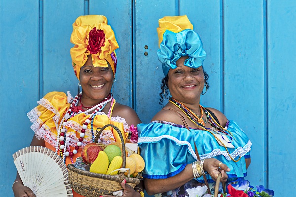 Portrait of happy Cuban women standing against blue wooden wall. Smiling mature women are in traditional dresses. They are with fruit basket and hand fan.