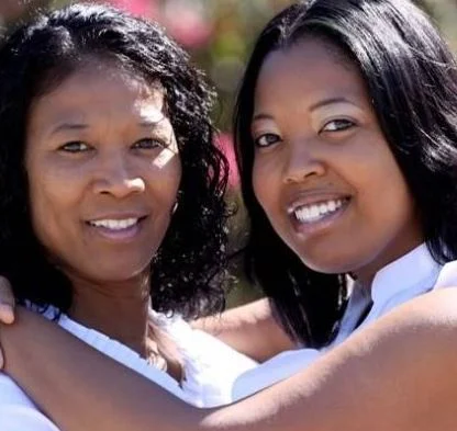 Two women smiling outdoors, one embracing the other with both arms.