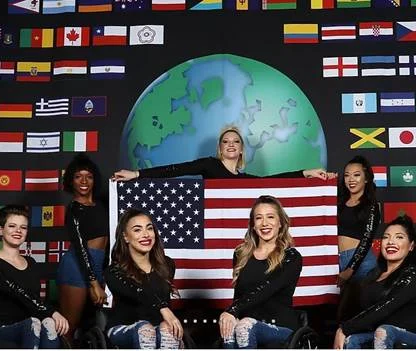 A group of nine women in black outfits pose with a U.S. flag. A backdrop shows various international flags and a globe.