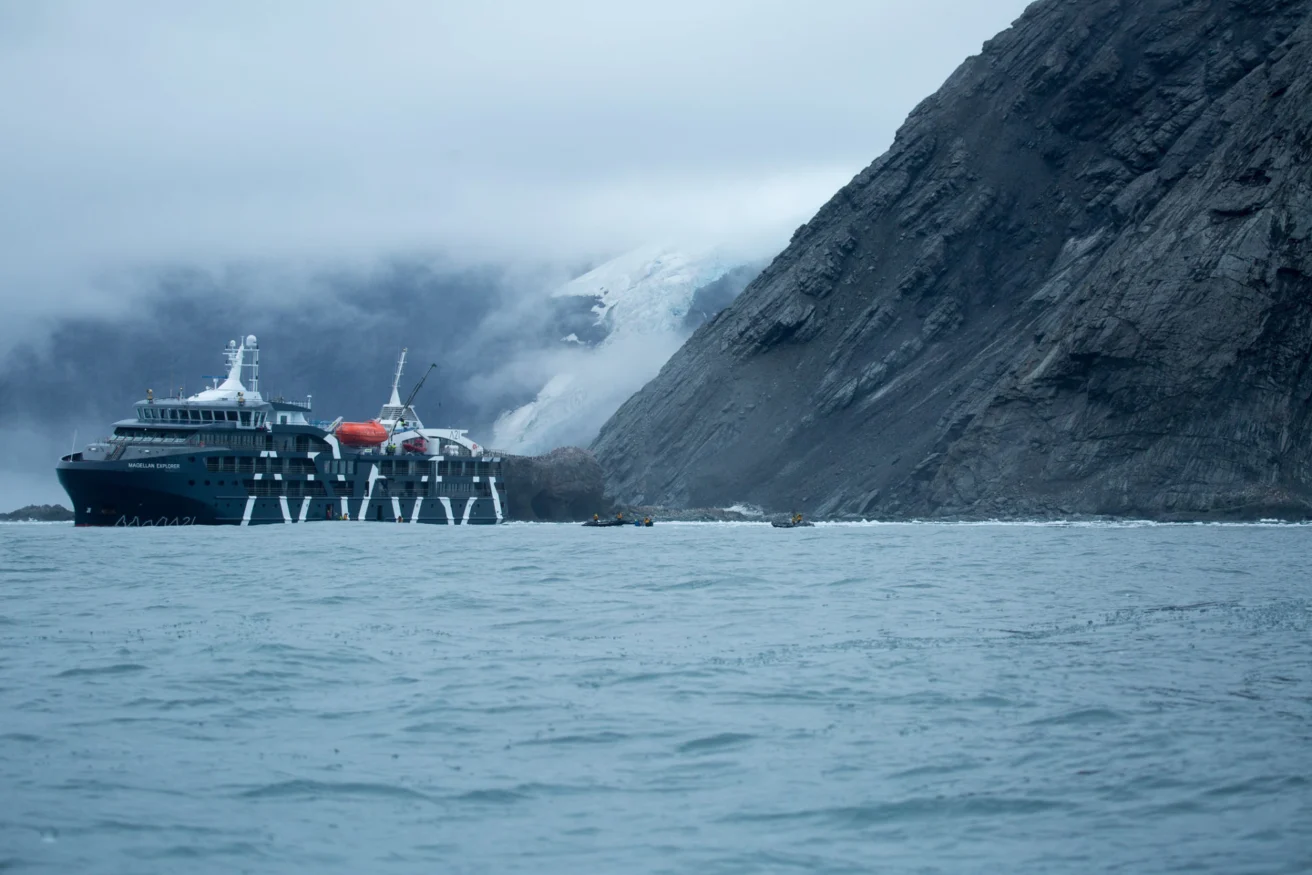 A cruise ship is anchored near a rocky coastline with a glacier in the background, under overcast skies.