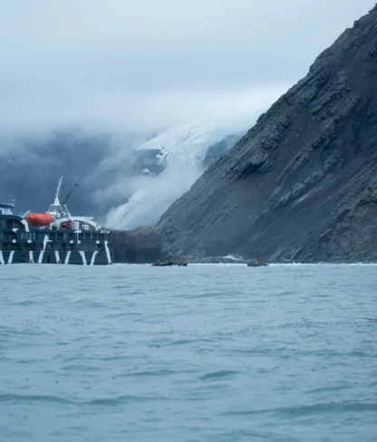 A cruise ship is anchored near a rocky coastline with a glacier in the background, under overcast skies.