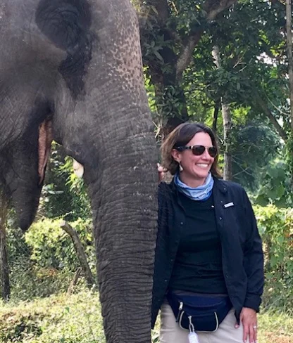 A person stands beside an elephant, smiling, in an outdoor setting with trees in the background.