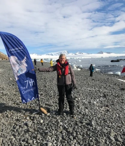 Person standing on a rocky shore in Antarctica, holding a flag that reads "The 7th Continent." Snowy mountains and glaciers are visible in the background.