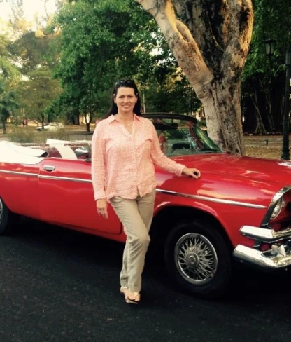 A woman stands next to a vintage red convertible car parked on a tree-lined road.