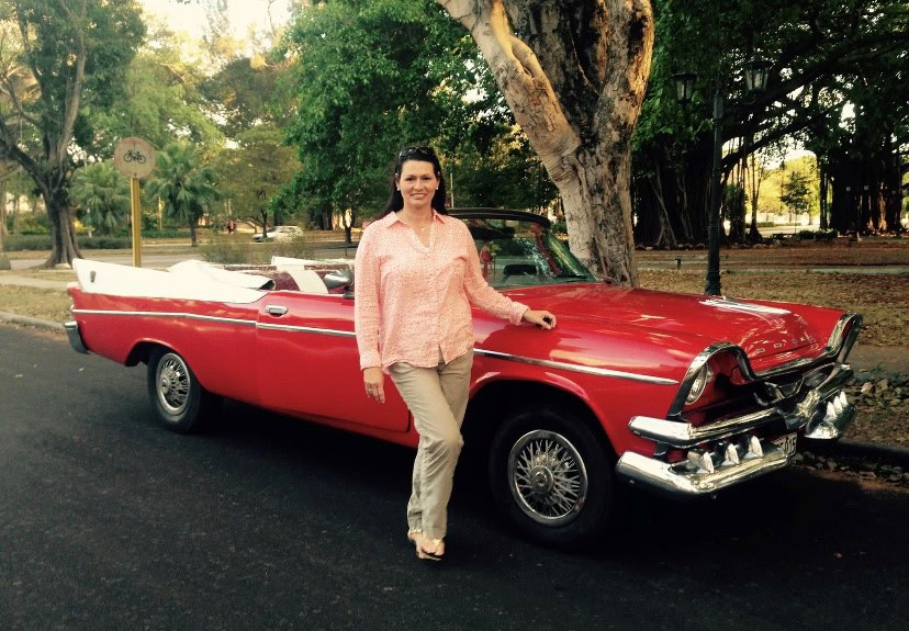 A woman stands next to a vintage red convertible car parked on a tree-lined road.