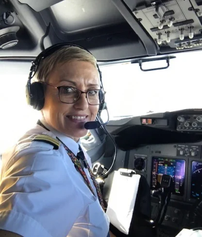 A pilot with short hair and glasses is smiling while seated in an aircraft cockpit, wearing a headset and uniform.