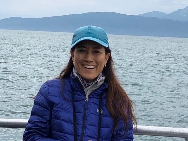 A person wearing a blue jacket and cap stands smiling in front of a body of water with mountains in the background.