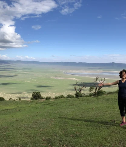 Person standing with arms outstretched on a grassy hillside overlooking a vast green valley under a cloudy sky.