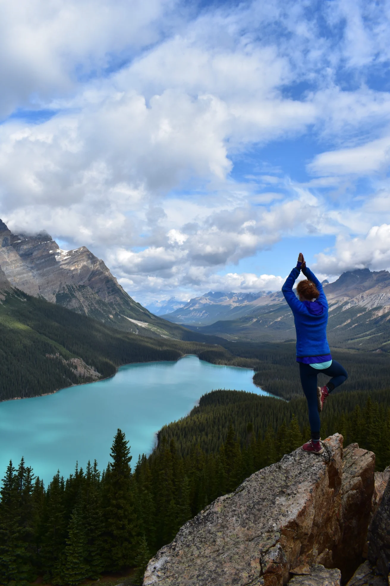 Person in a blue jacket doing a yoga pose on a rocky cliff overlooking a scenic lake and mountains under a cloudy sky.