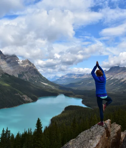 Person in a blue jacket doing a yoga pose on a rocky cliff overlooking a scenic lake and mountains under a cloudy sky.