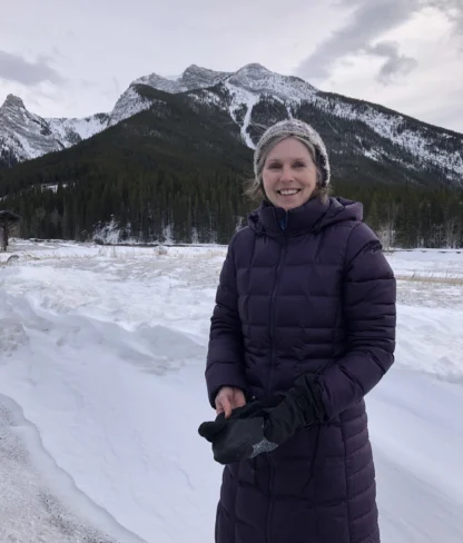 A person in a purple coat stands in a snowy landscape with mountains in the background.