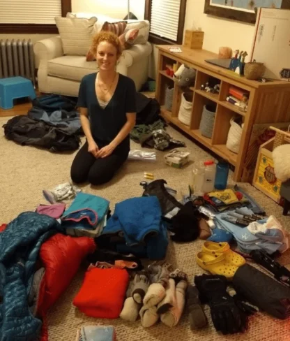 Person kneeling on a carpet surrounded by assorted camping gear, including sleeping bags, clothing, shoes, and supplies, in a living room setting.