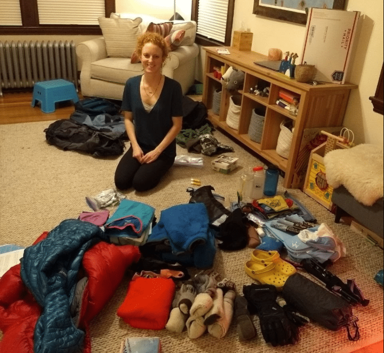 Person kneeling on a carpet surrounded by assorted camping gear, including sleeping bags, clothing, shoes, and supplies, in a living room setting.