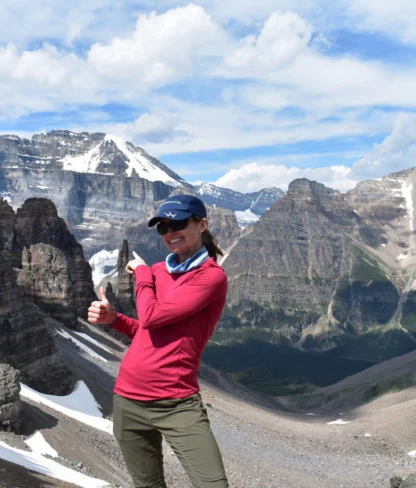 A person in a red shirt poses with a thumbs-up on a rocky mountain landscape under a blue sky with clouds. Snow-capped peaks are visible in the background.