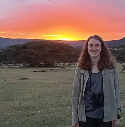 A person stands in a field at sunset with binoculars around their neck. Trees and hills are in the background under a colorful sky.