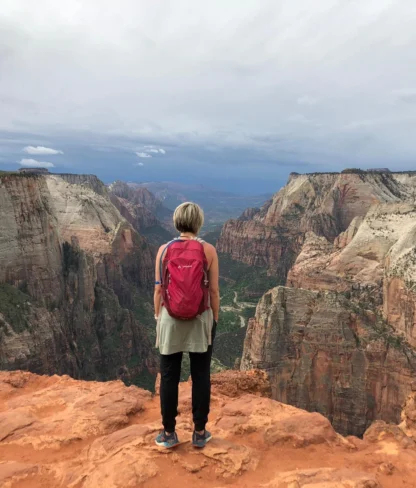 A person with a red backpack stands at the edge of a cliff, overlooking a vast canyon under a cloudy sky.