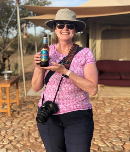 A person in a hat and sunglasses holds a bottle of beer, standing outside a tent on a sunny day, with a camera around their neck.