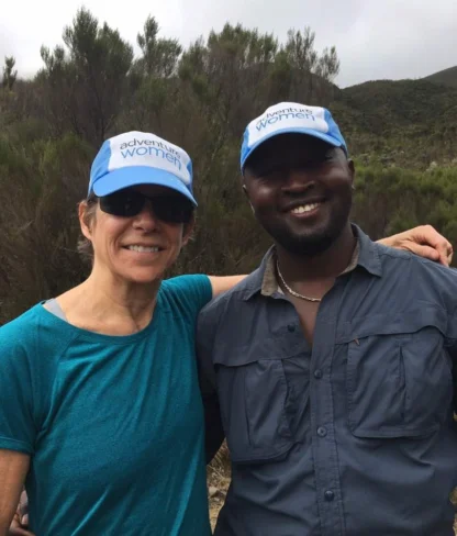 Two people wearing blue "adventure women" hats and casual outdoor clothing pose together, smiling, in a natural setting with green foliage in the background.