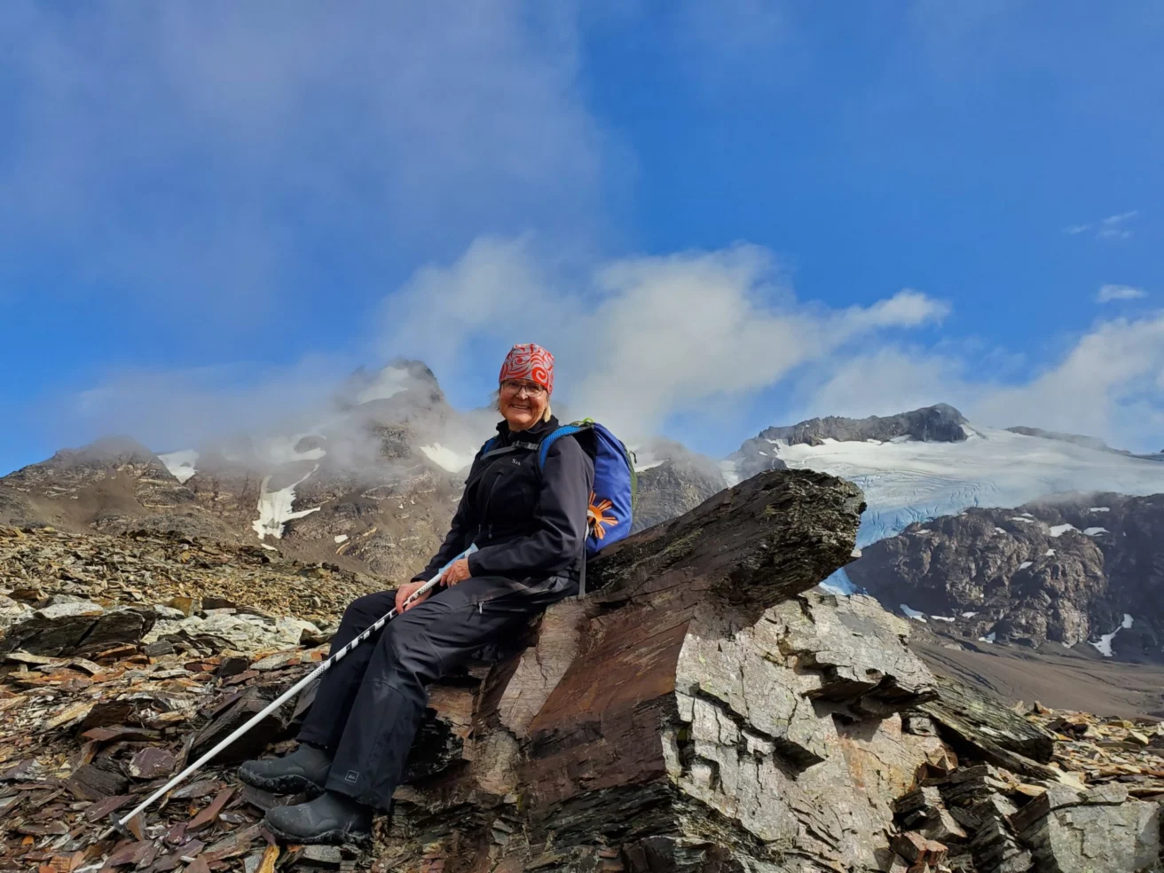 A person wearing hiking gear and a red bandana sits on a rock with a blue backpack, surrounded by rocky terrain and snow-capped mountains under a blue sky.