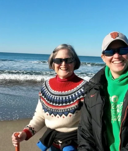 Two people in sunglasses and warm clothing smile on a sunny beach with waves in the background.