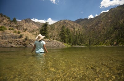 A person wearing a hat stands in clear, shallow water, facing a landscape of trees and mountains under a blue sky.