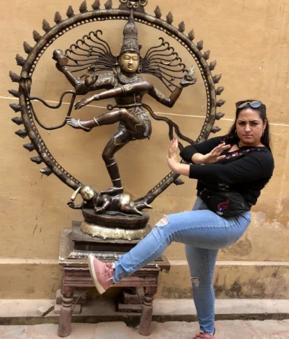 A woman strikes a similar pose to a bronze Nataraja statue, both with raised arms and legs. The statue stands on a wooden pedestal against a beige wall.