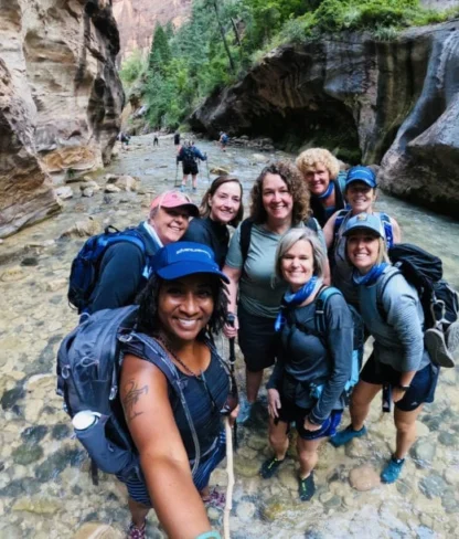 A group of hikers stand in a shallow river surrounded by rocky cliffs and trees, smiling for a photo.