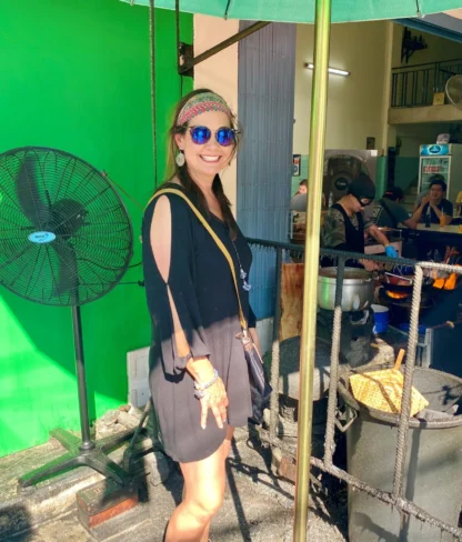 A person in sunglasses and a black dress stands smiling near a sidewalk food stall with a large fan nearby. Other people are eating in the background.