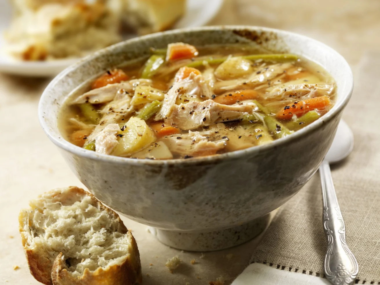 A bowl of chicken soup with vegetables, including carrots and celery, is on a table with a metal spoon and a piece of bread.