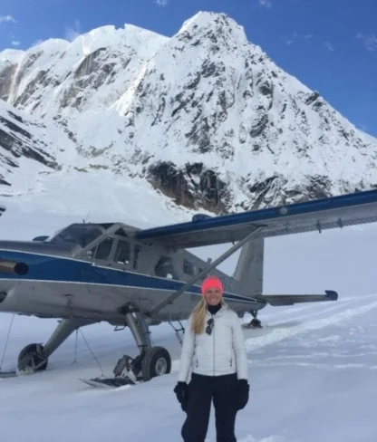 A person in winter clothing stands in the snow next to a small airplane, with a large snow-covered mountain in the background.