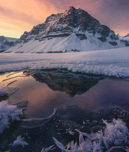 Snow-covered mountain under a colorful sky is reflected in a partially frozen lake with ice crystals along the edges.