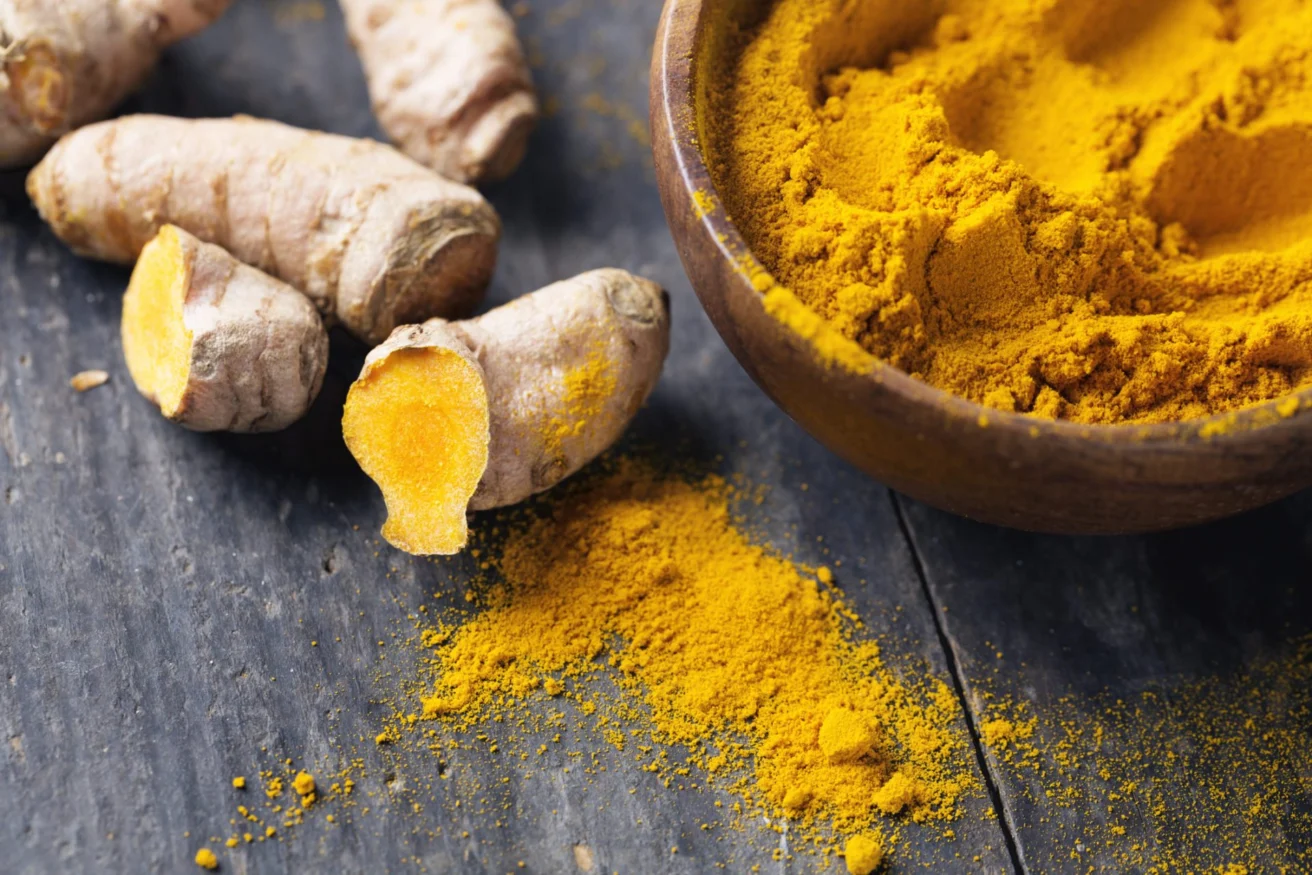 Turmeric roots and ground turmeric in a wooden bowl are placed on a dark wooden surface. Turmeric powder is scattered nearby.