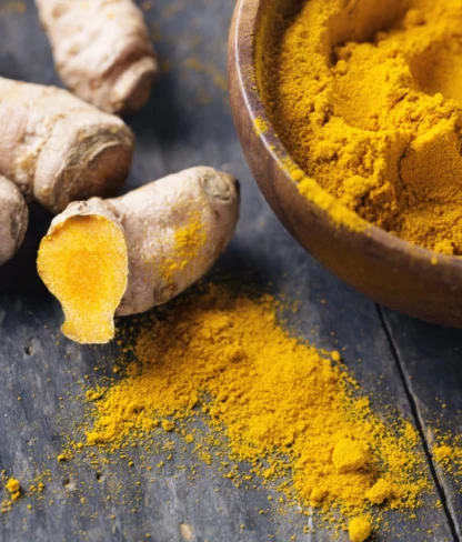 Turmeric roots and ground turmeric in a wooden bowl are placed on a dark wooden surface. Turmeric powder is scattered nearby.