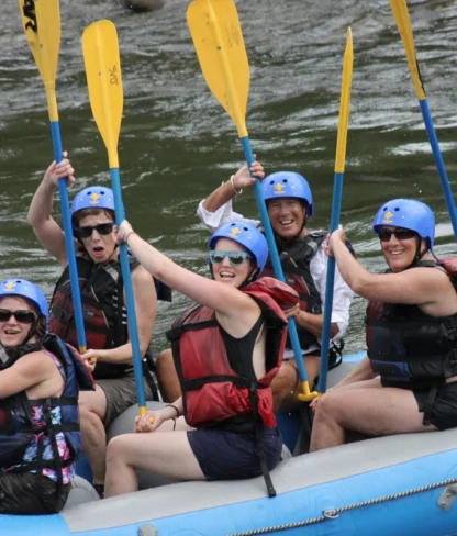 Group of people wearing helmets and life jackets holding paddles while sitting in a blue raft on a river.