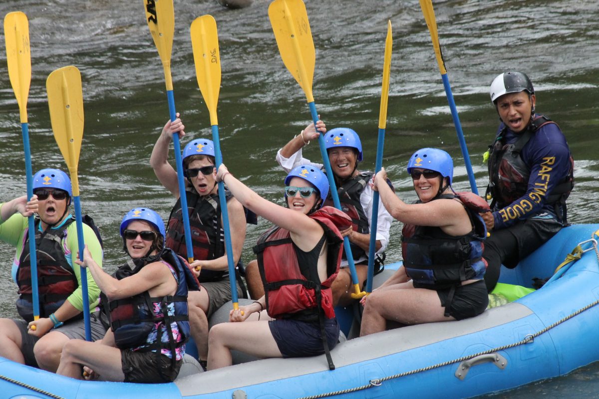 Group of people wearing helmets and life jackets holding paddles while sitting in a blue raft on a river.