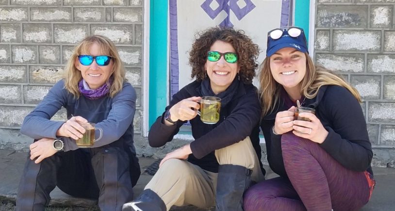 Three women laughing and smiling sitting against a building before hiking.