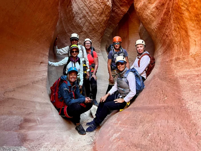 A group of seven people wearing helmets and backpacks stand in a narrow, red rock canyon.