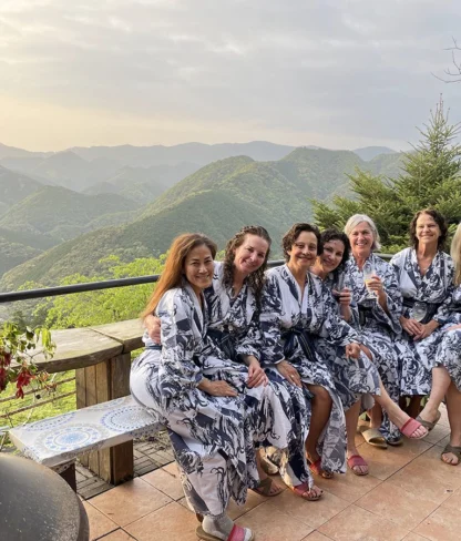 Seven women in matching robes sit on a bench overlooking a lush, mountainous landscape.