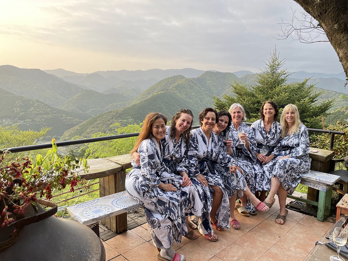 Seven women in matching robes sit on a bench overlooking a lush, mountainous landscape.