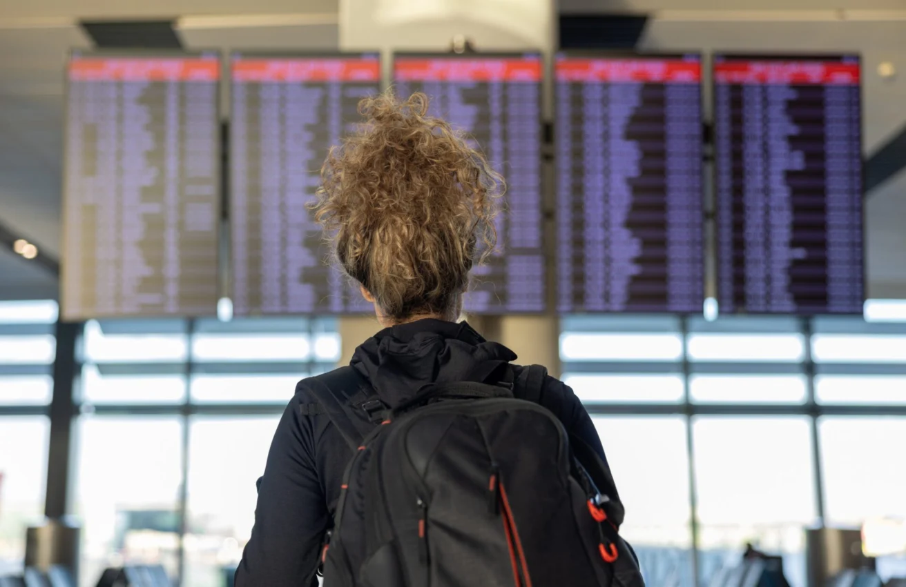 A person with curly hair and a backpack stands facing an airport departures board showing numerous flights.