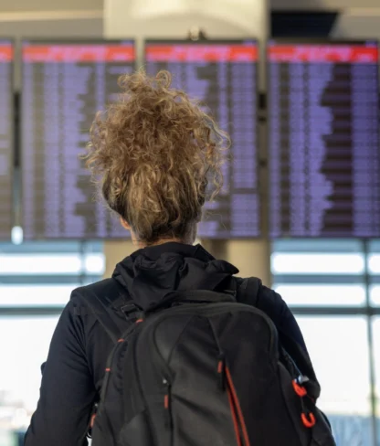 A person with curly hair and a backpack stands facing an airport departures board showing numerous flights.