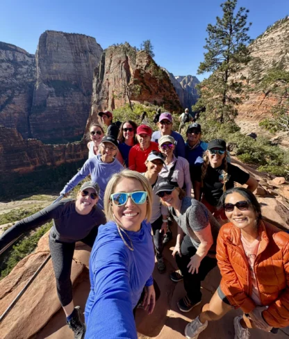 A group of people taking a selfie on a mountain trail with a scenic canyon view in the background.