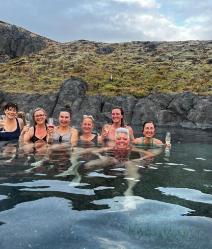 A group of people relax and enjoy drinks in a natural hot spring surrounded by rocky terrain.